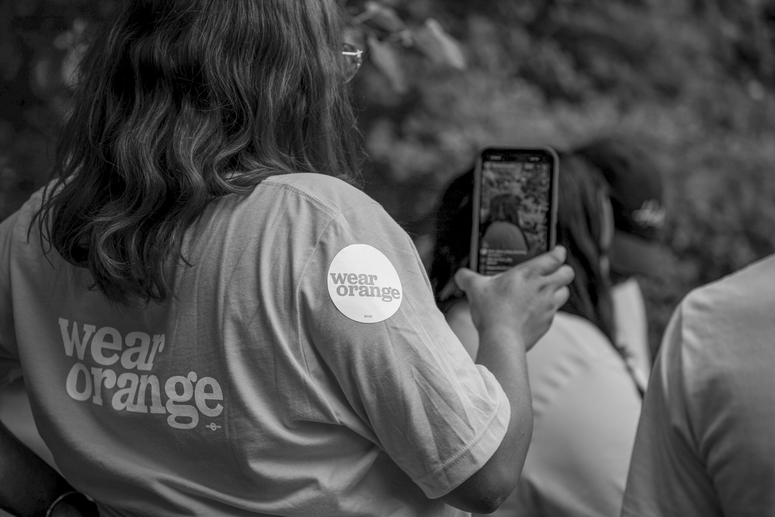 A black-and-white image of a woman wearing a wear orange t-shirt and recording what appears to be a community event on her phone.